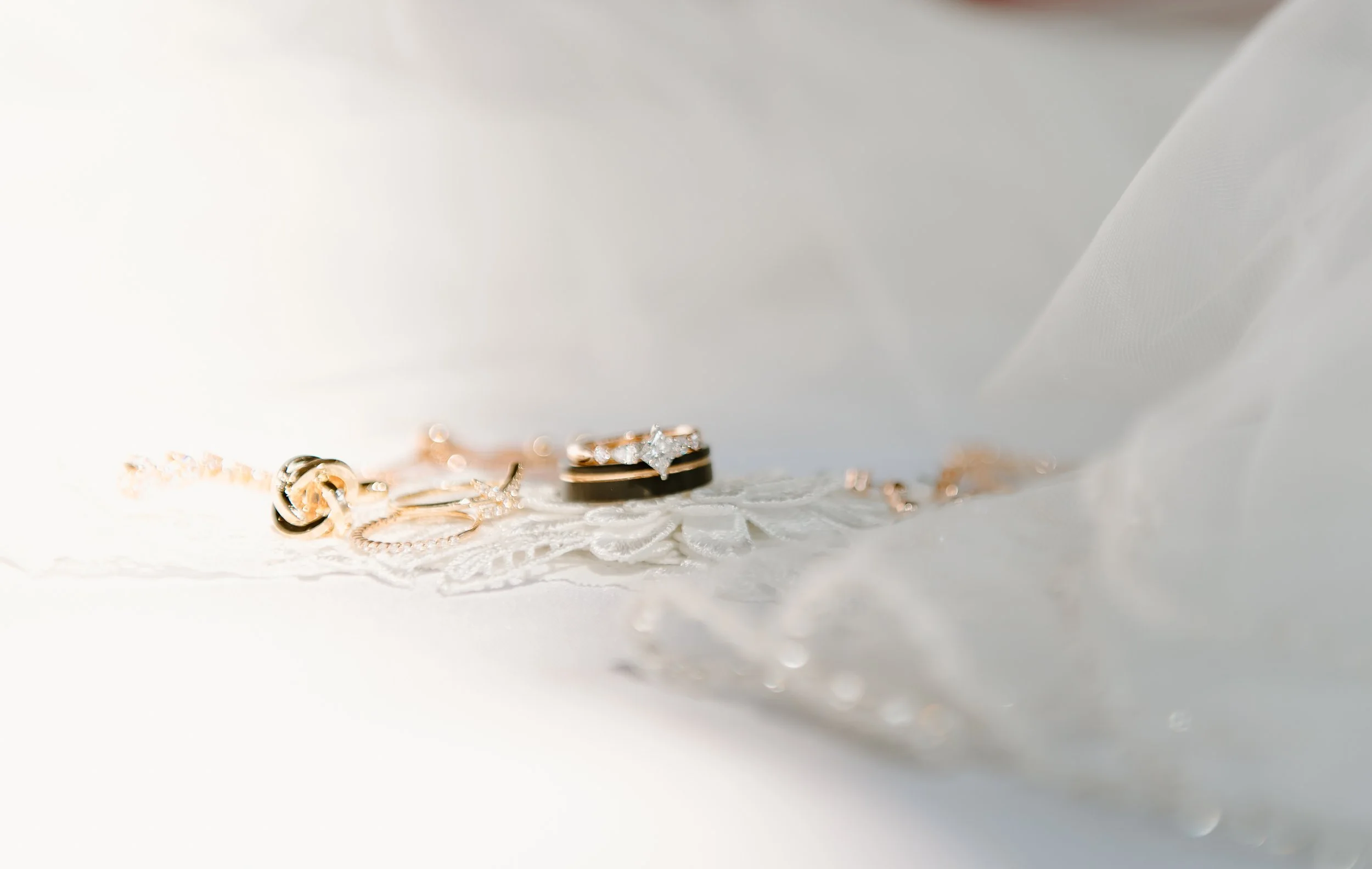 Close-up of gold and silver jewelry, including rings and a bracelet, on a white lace cloth, with a soft white background.