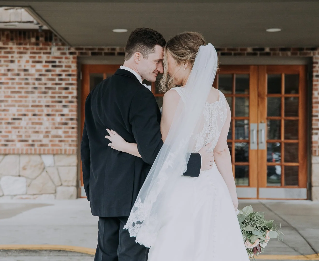 A bride and groom embrace each other closely outside a building with a brick wall and wooden double doors in the background.