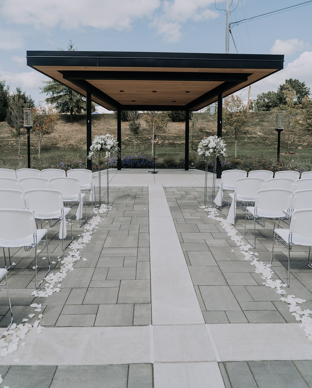 Outdoor wedding ceremony setup with white chairs, floral arrangements, and a black and wood canopy under a partly cloudy sky.