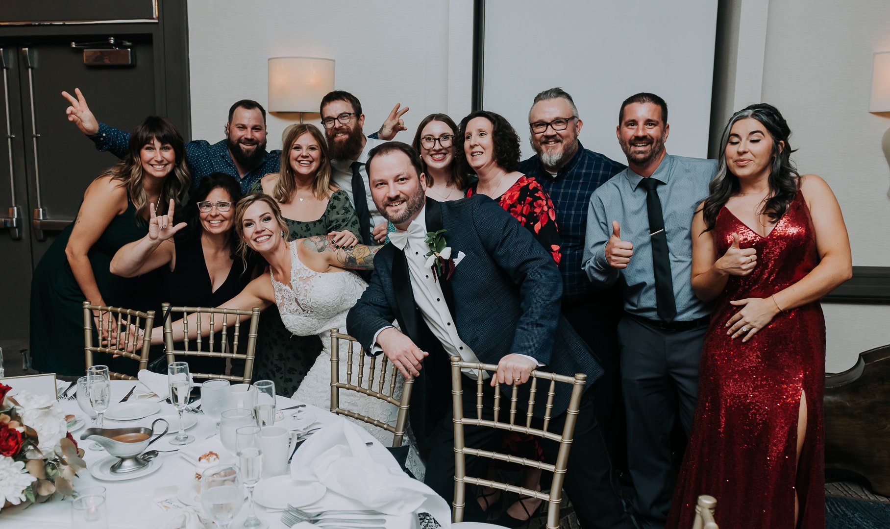 A group of people at a wedding celebration, including the bride and groom, posing cheerfully at a banquet table.