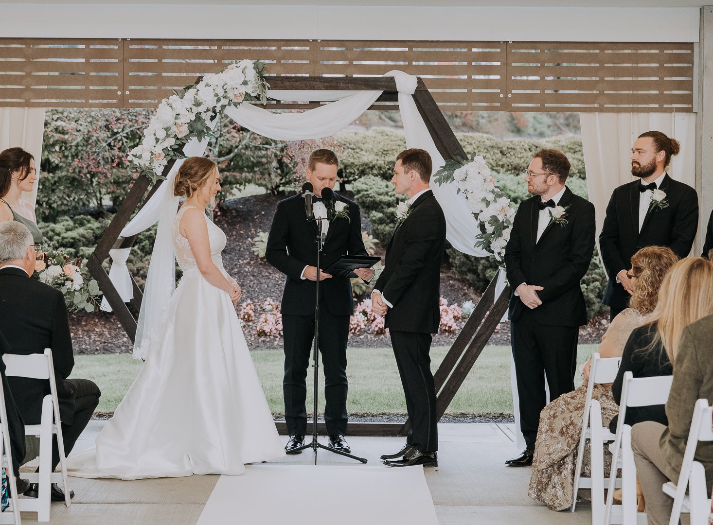 A wedding ceremony with a bride and groom standing under a decorated arch, exchanging vows in an outdoor setting, with guests seated on either side.