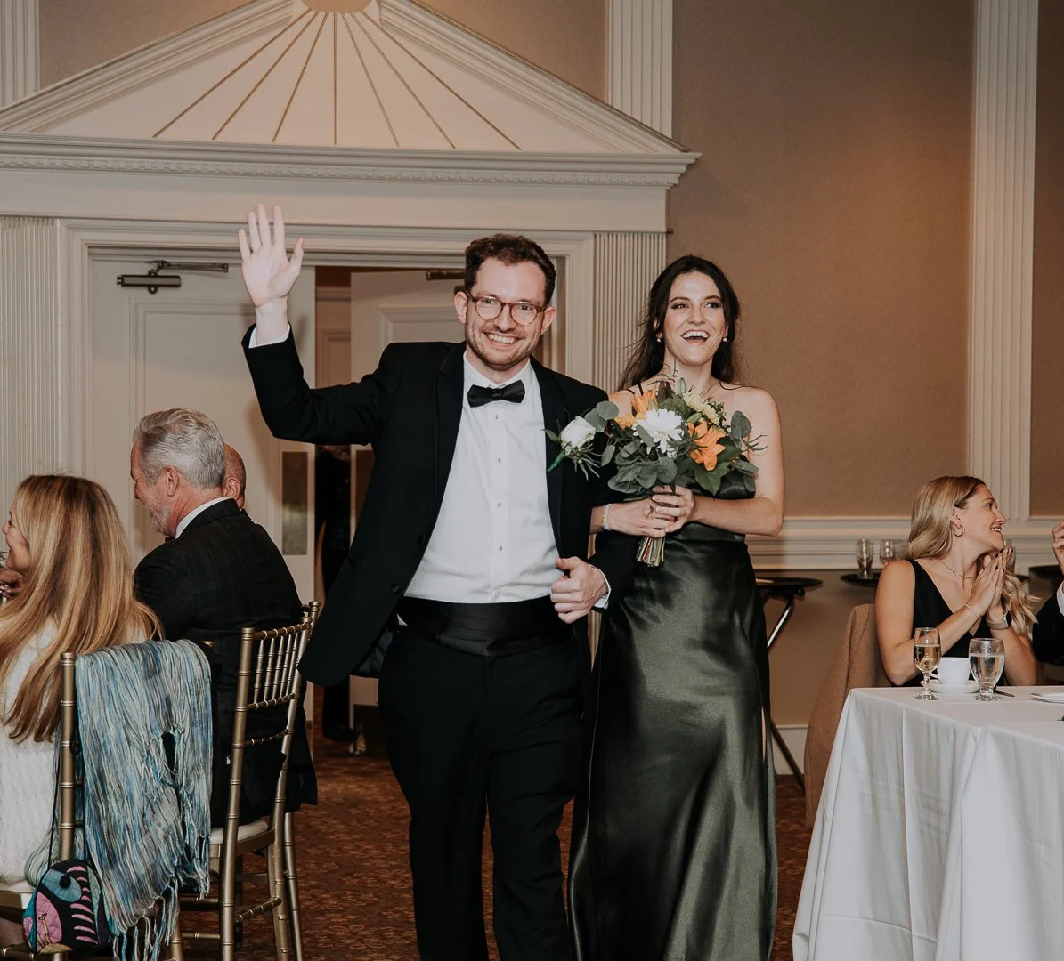 A man and woman at a formal event, possibly a wedding reception, with the man waving and smiling, and the woman holding a bouquet of flowers and smiling. Other guests are seated at tables in the background.