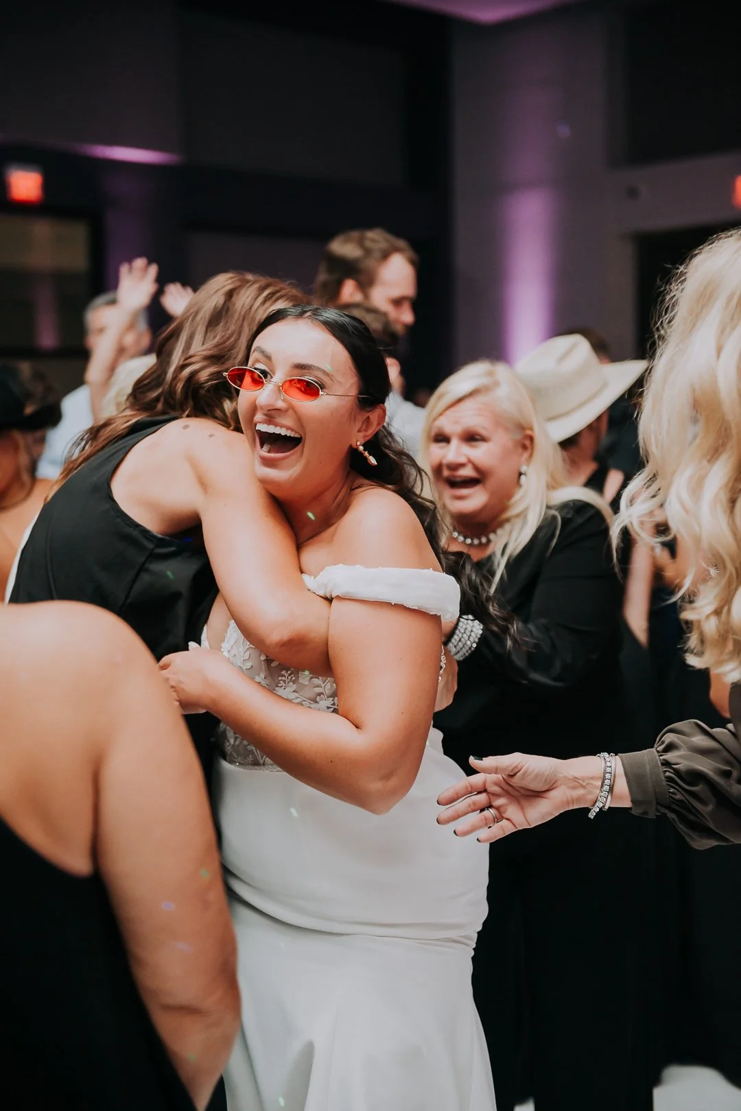 People dancing and celebrating at a wedding reception, with a woman in a white dress and red sunglasses smiling and hugging a man.