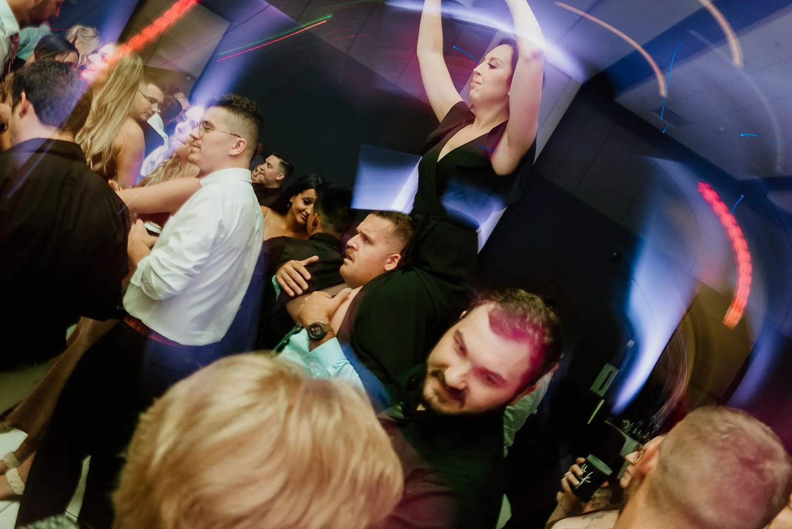 A crowded party scene with people dancing and socializing, including a woman sitting on a man's shoulders and others engaged in conversations under colorful lighting.