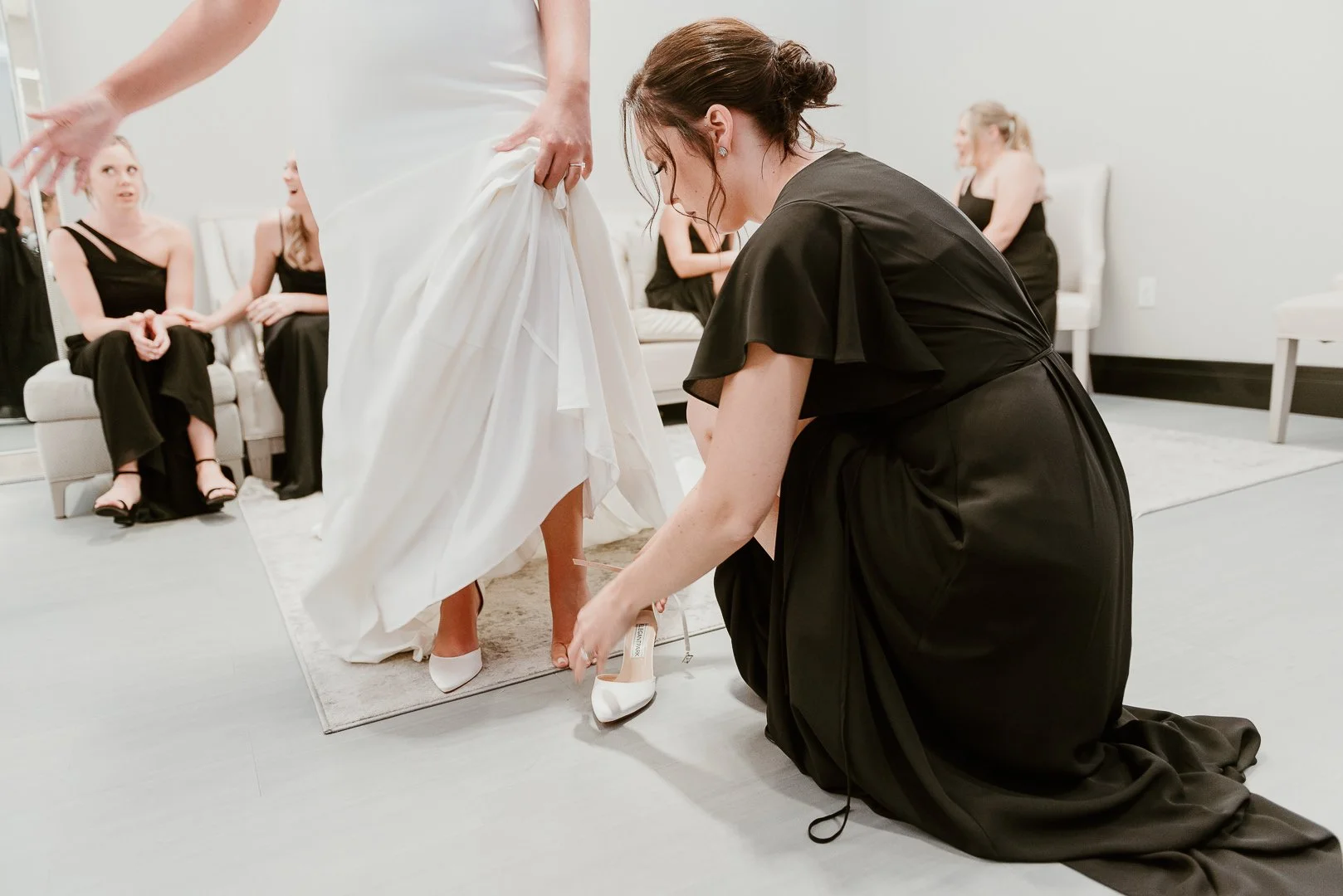 Women dressing in black and white dresses, with one woman kneeling and adjusting a white high heel shoe on another woman's foot while other women sit on a couch in the background.