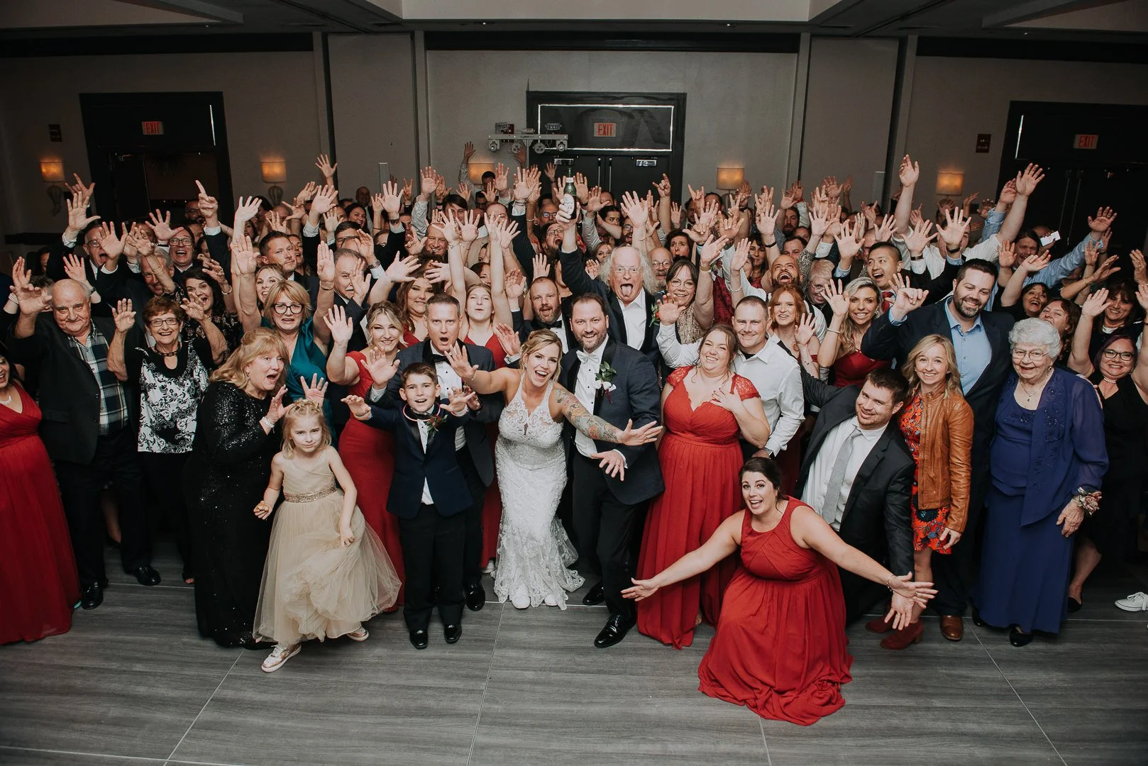 Large group of people at a wedding celebration, many with hands raised, smiling, and cheerful. The bride and groom are in the front, surrounded by family and friends in formal and semi-formal attire.
