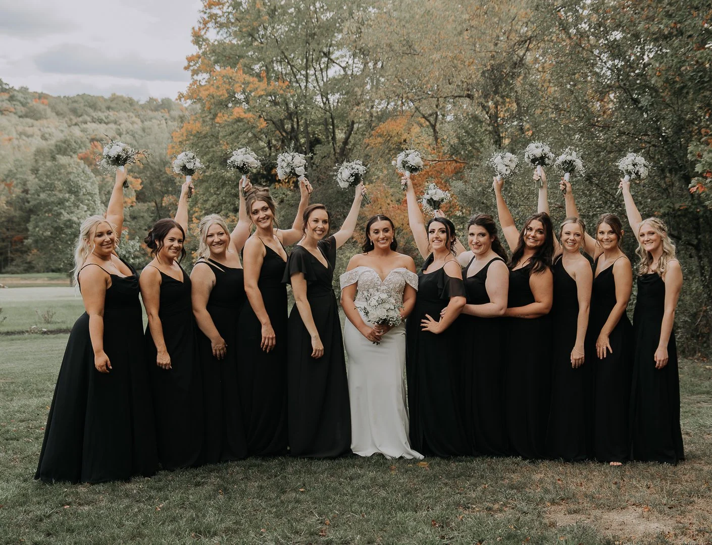 A bride in a white wedding dress standing with her bridesmaids dressed in black dresses outdoors, with some bridesmaids holding up bouquets in celebration.