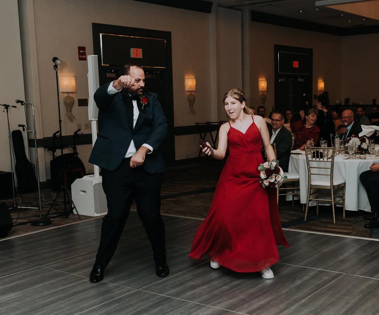 Two people dancing at a wedding reception, a man in a tuxedo with a red boutonniere and a woman in a red dress holding a bouquet. Guests seated at tables in the background are watching and smiling.
