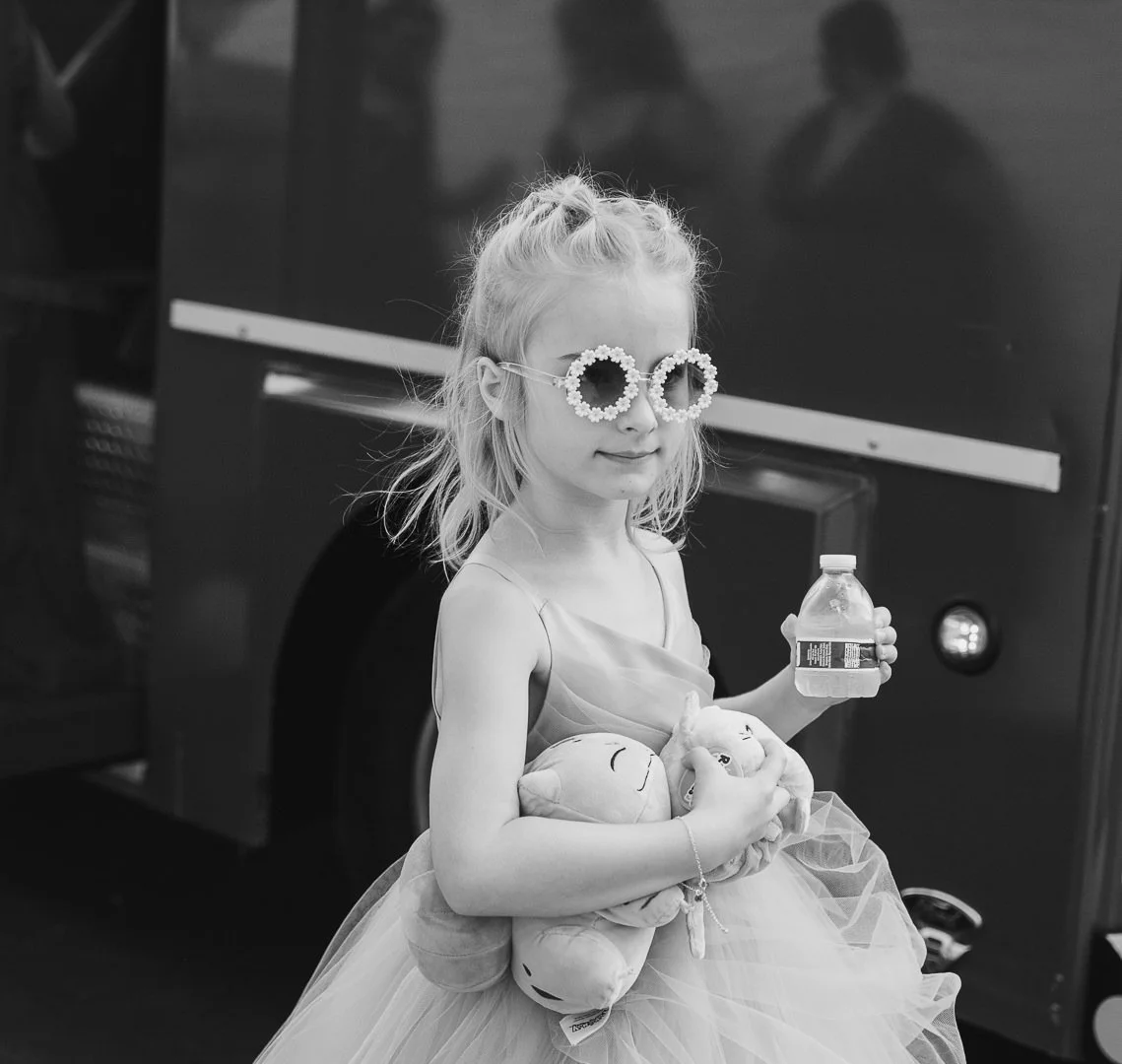 A young girl with blonde hair wearing sunglasses with a flower design, holding a bottle of water and plush dolls, standing in front of a vehicle.