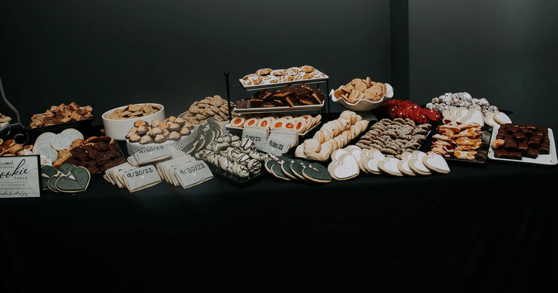 A table filled with various cookies and sweet treats, decorated with heart-shaped cookies and signs, set against a dark background.