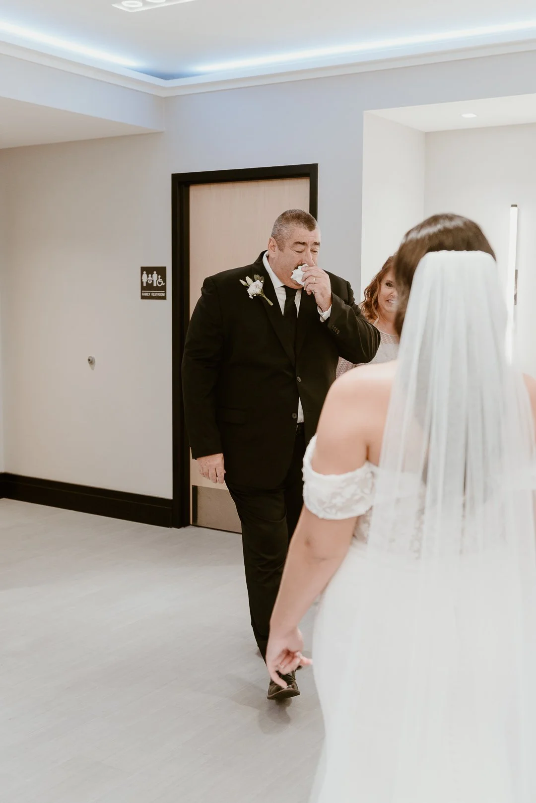 A man in a black suit and a woman in a white wedding dress are in an indoor hallway, with the man wiping his nose with a tissue and smiling, and another woman smiling in the background.