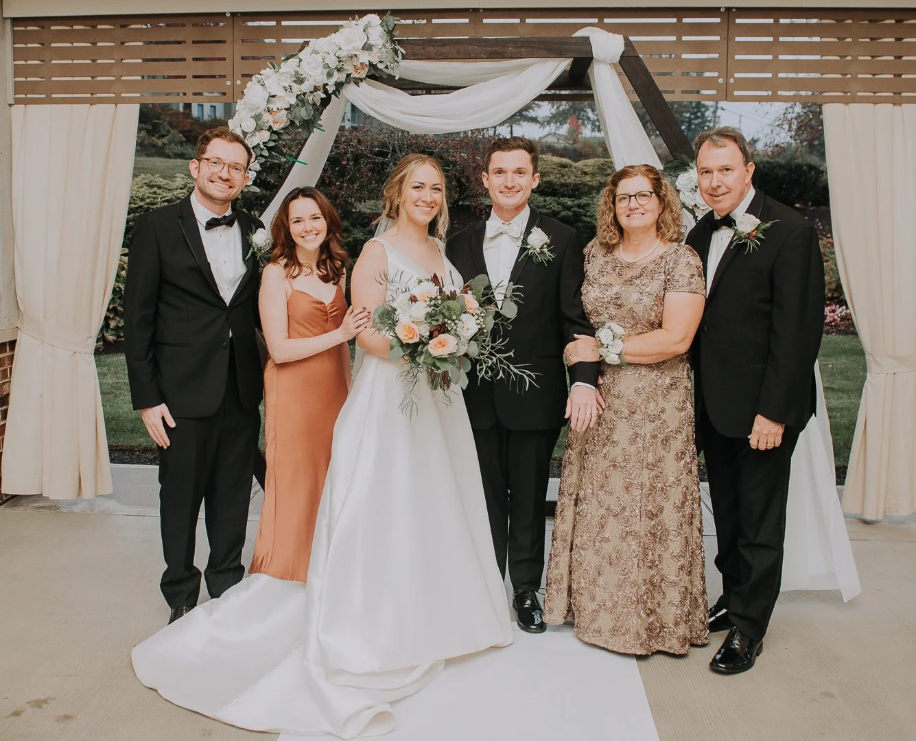 Group of seven people, including bride and groom, posing under a wedding arch with flowers.