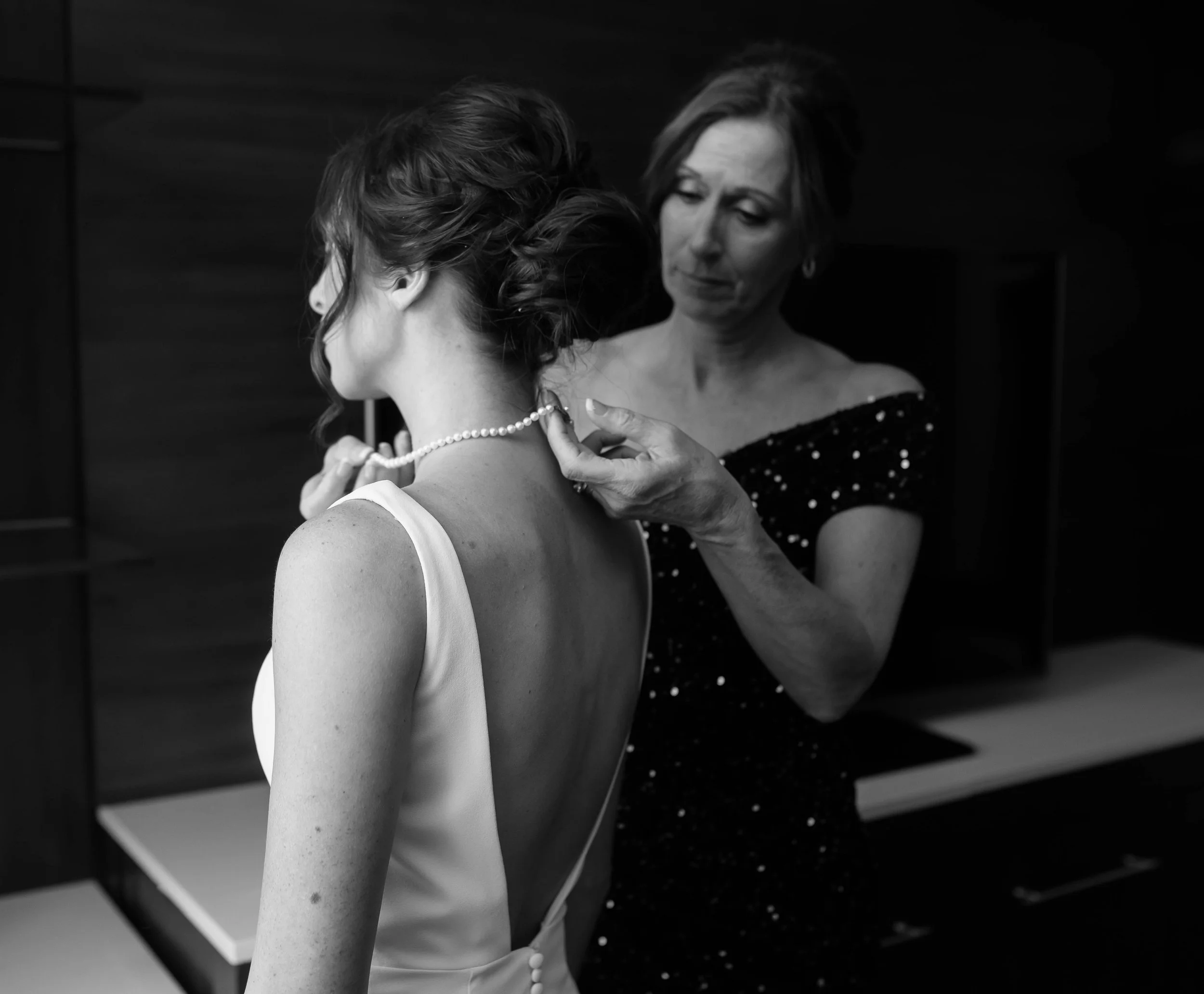 A woman is helping another woman with a pearl necklace; the woman being helped is in a white dress, while the helper is in a dark, sparkly dress, both in an indoor setting.