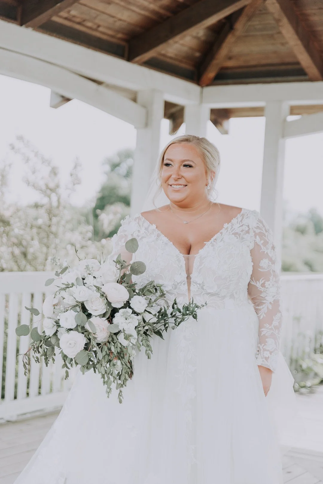 A bride in a white wedding dress holding a bouquet of white flowers and greenery, smiling outside on a covered porch.