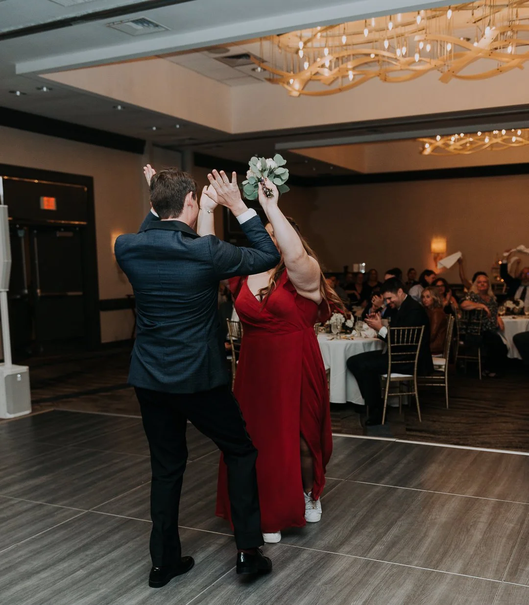A couple dancing at a wedding reception, with guests watching and seated at tables in the background.
