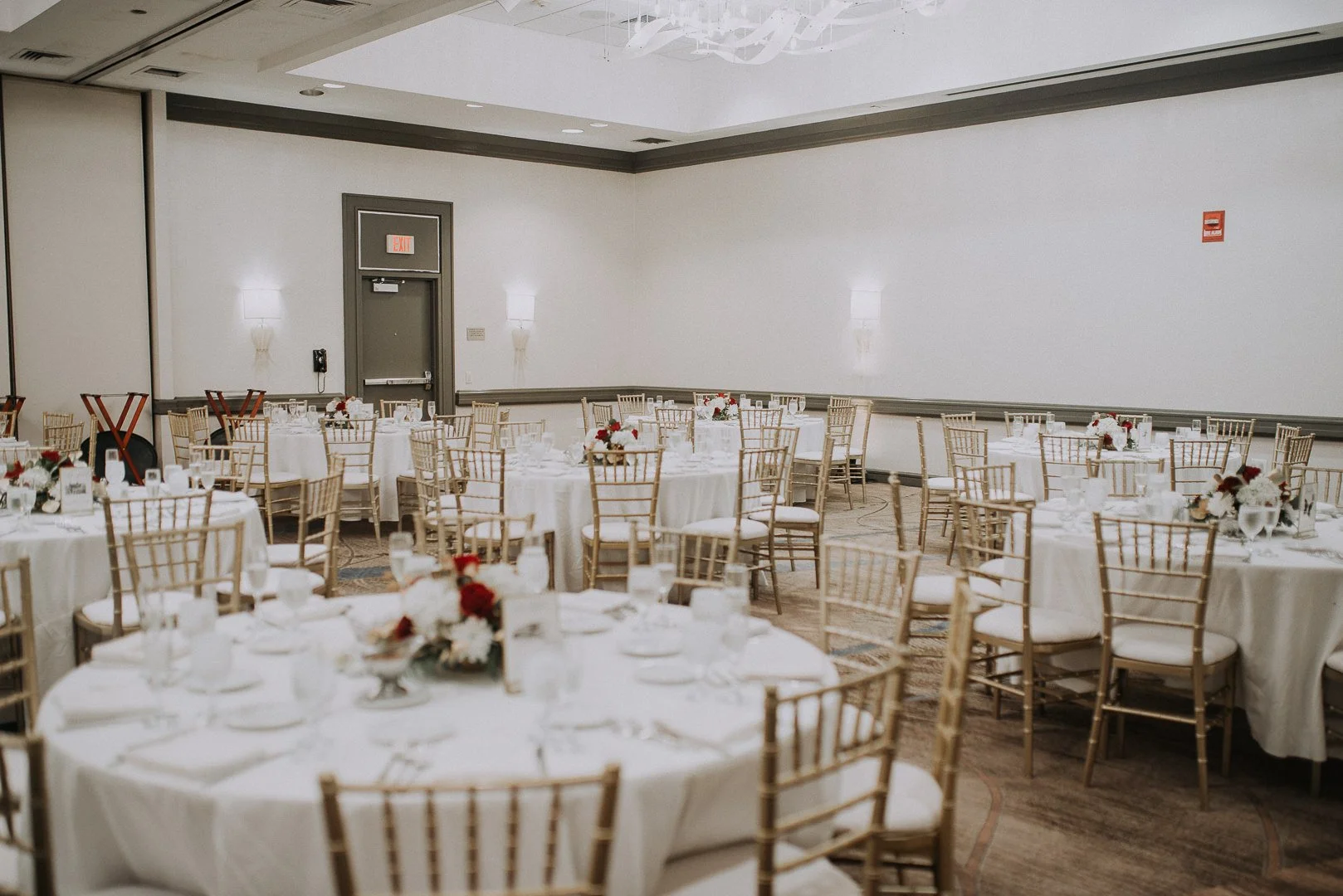 A banquet hall set up for a formal event with round tables covered in white tablecloths, decorated with floral centerpieces, and surrounded by gold Chiavari chairs with white cushions. The room has plain white walls, wall-mounted lights, and a gray d