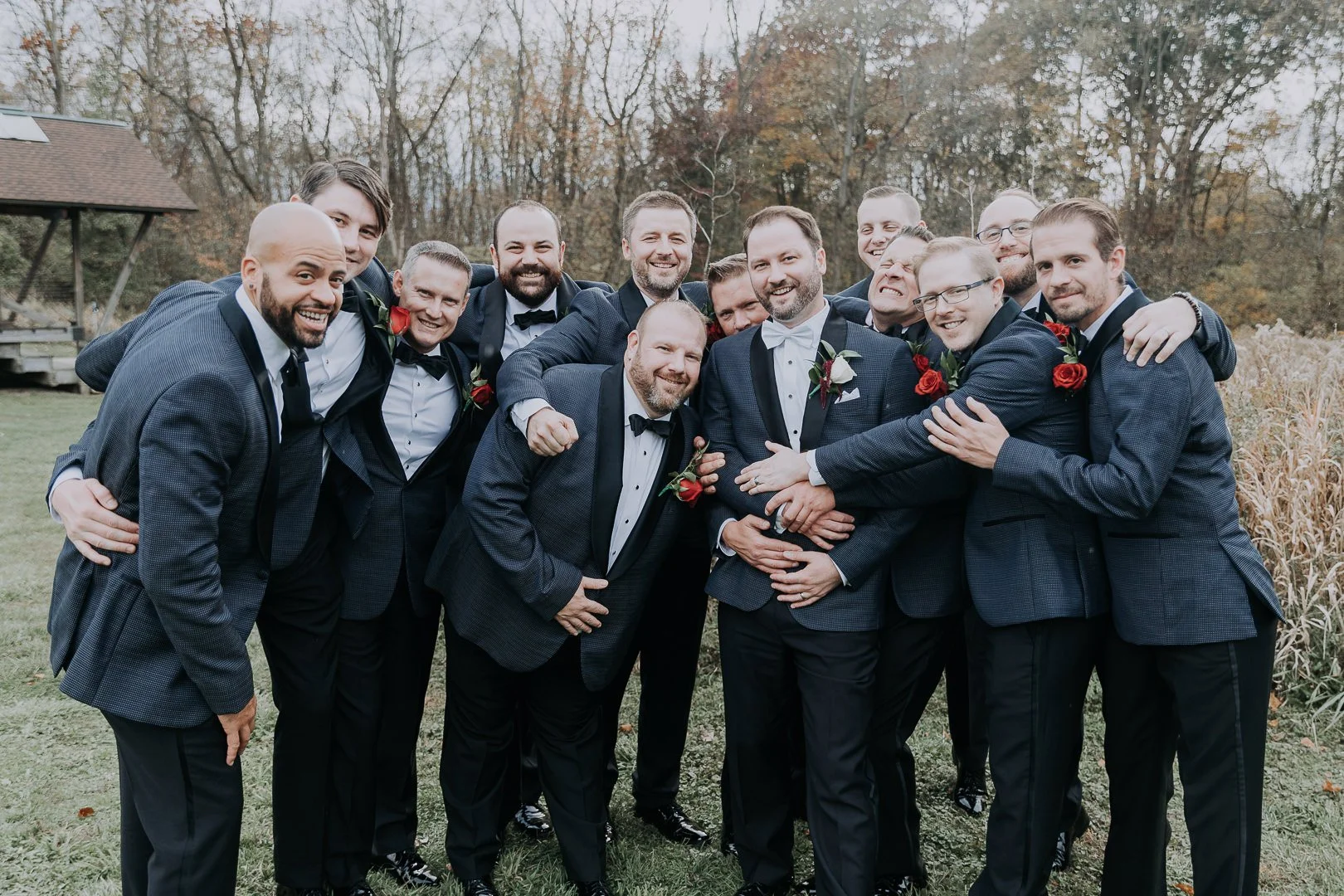 Group of men in tuxedos smiling and embracing outdoors during fall.