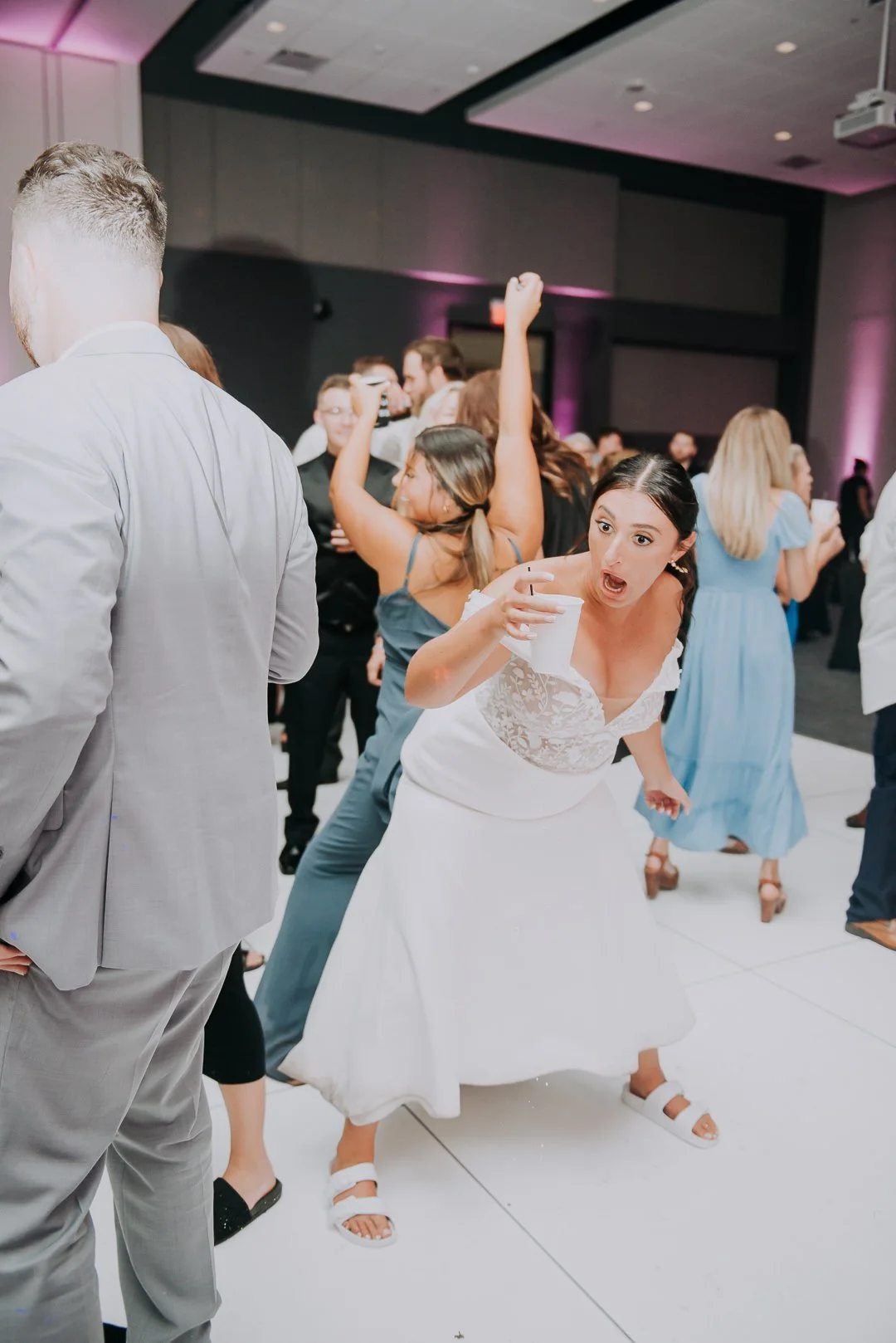 A woman in a white dress dances excitedly at a party, holding a white cup, surrounded by people dancing and enjoying the celebration.