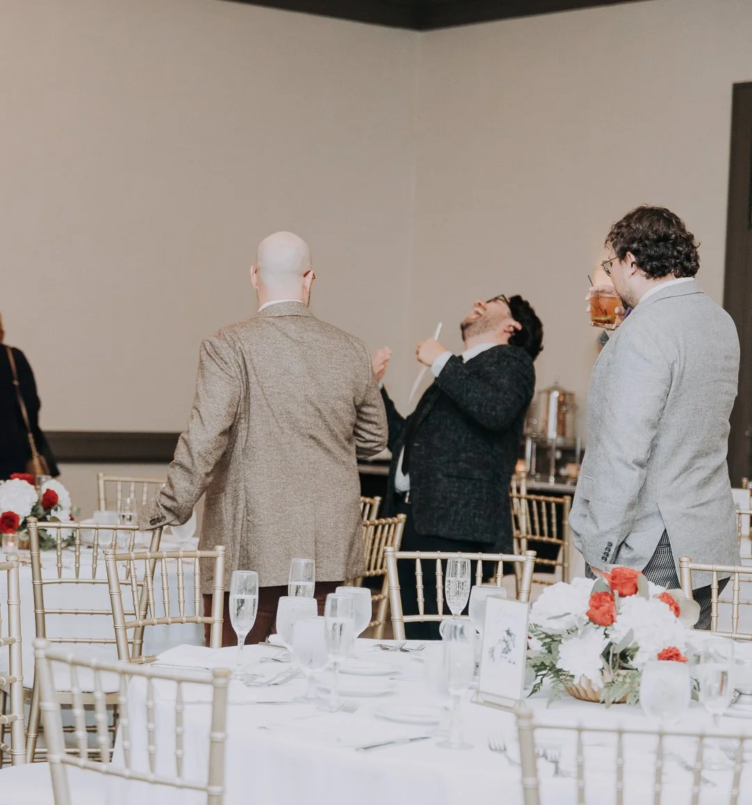 Three men in formal attire standing and laughing at a social gathering in a decorated banquet hall.