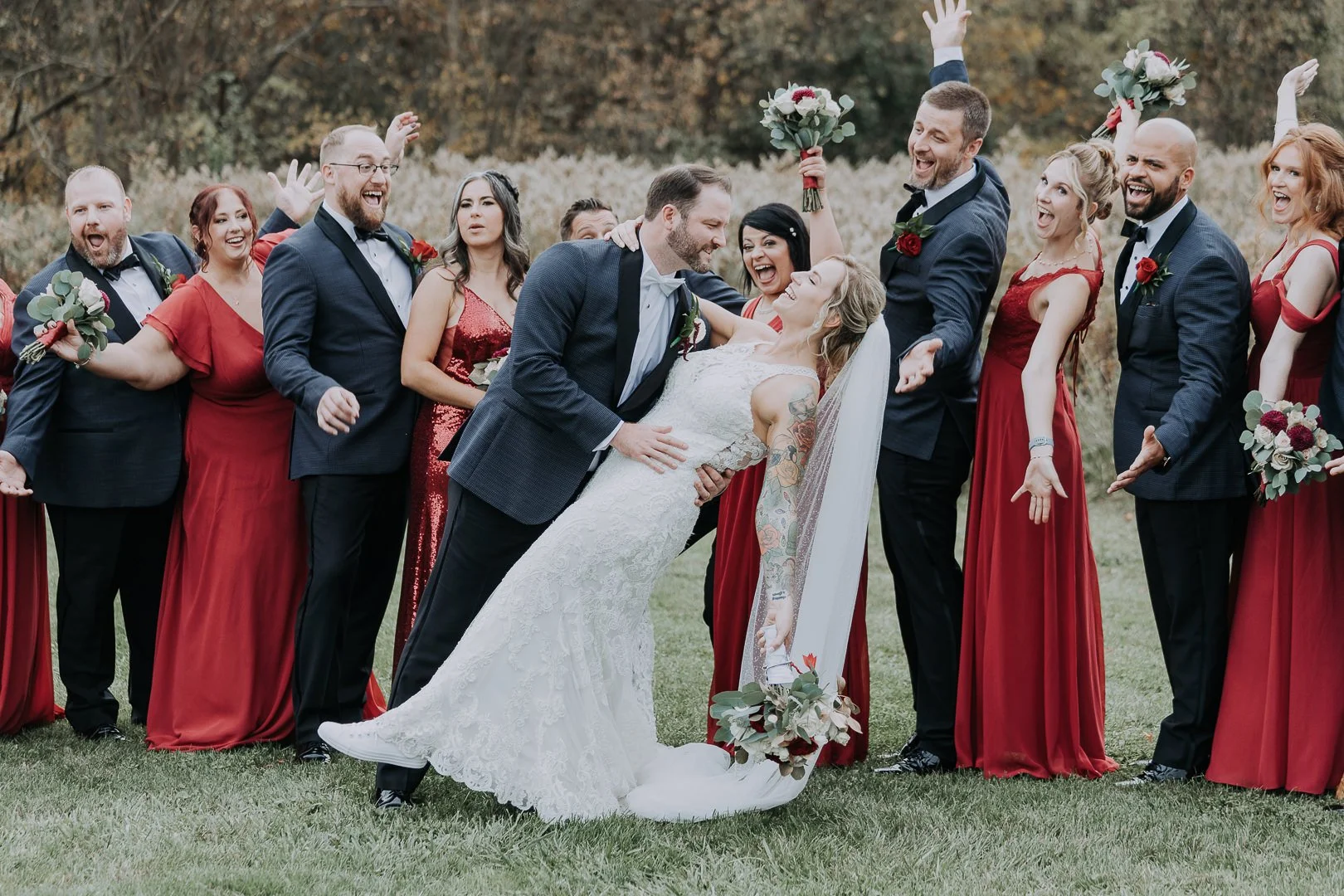 A wedding party outdoors with the bride and groom in the center. The groom is dipping the bride and they are smiling at each other. The bride holds a bouquet raised in the air, and the others around are dressed in formal attire, mostly red dresses an