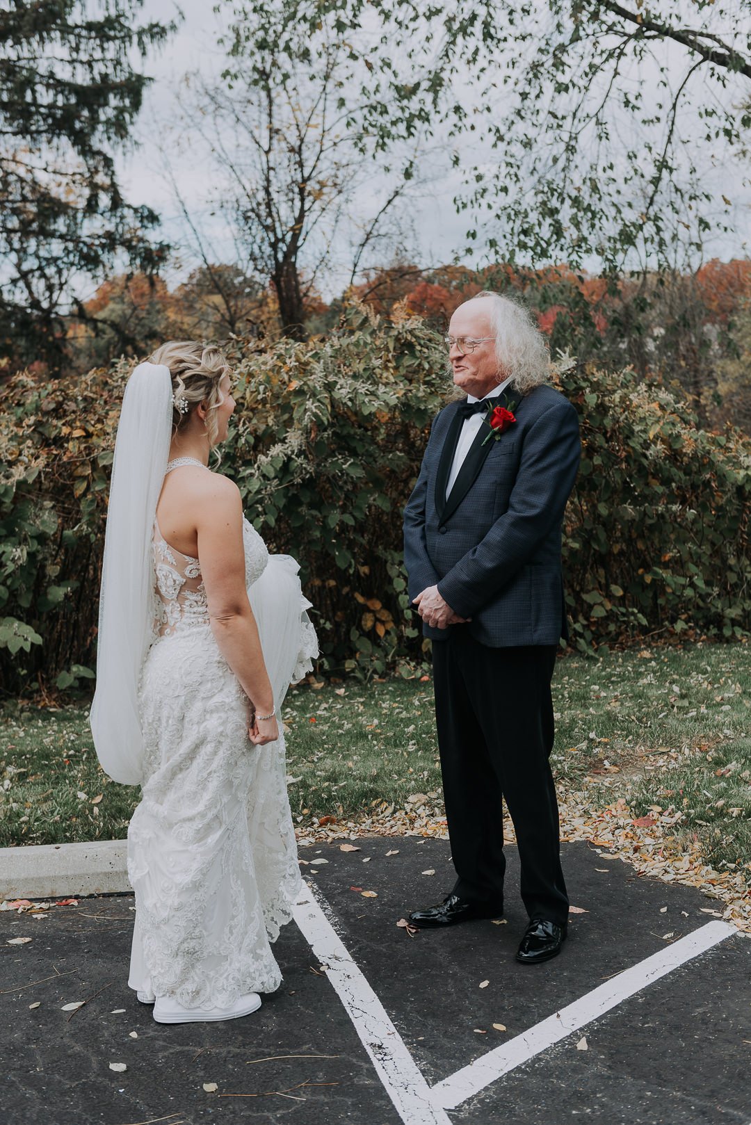 A bride and an elderly man are standing outdoors in a parking lot, facing each other, with autumn trees and bushes in the background. The bride is wearing a white lace wedding dress with a veil, and the man is dressed in a tuxedo with a red flower bo