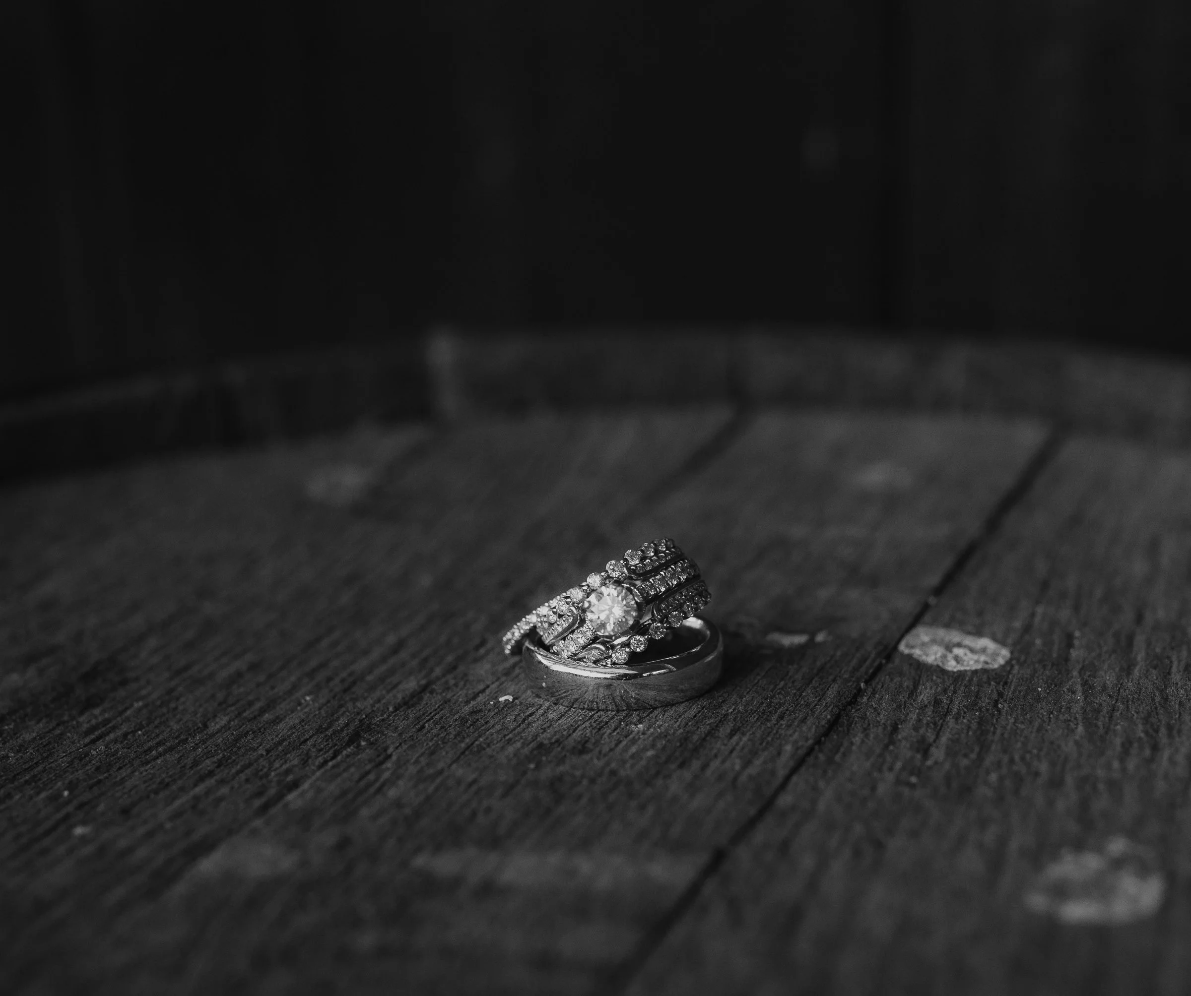 Black and white photo of rings, including a diamond engagement ring, placed on a wooden surface.