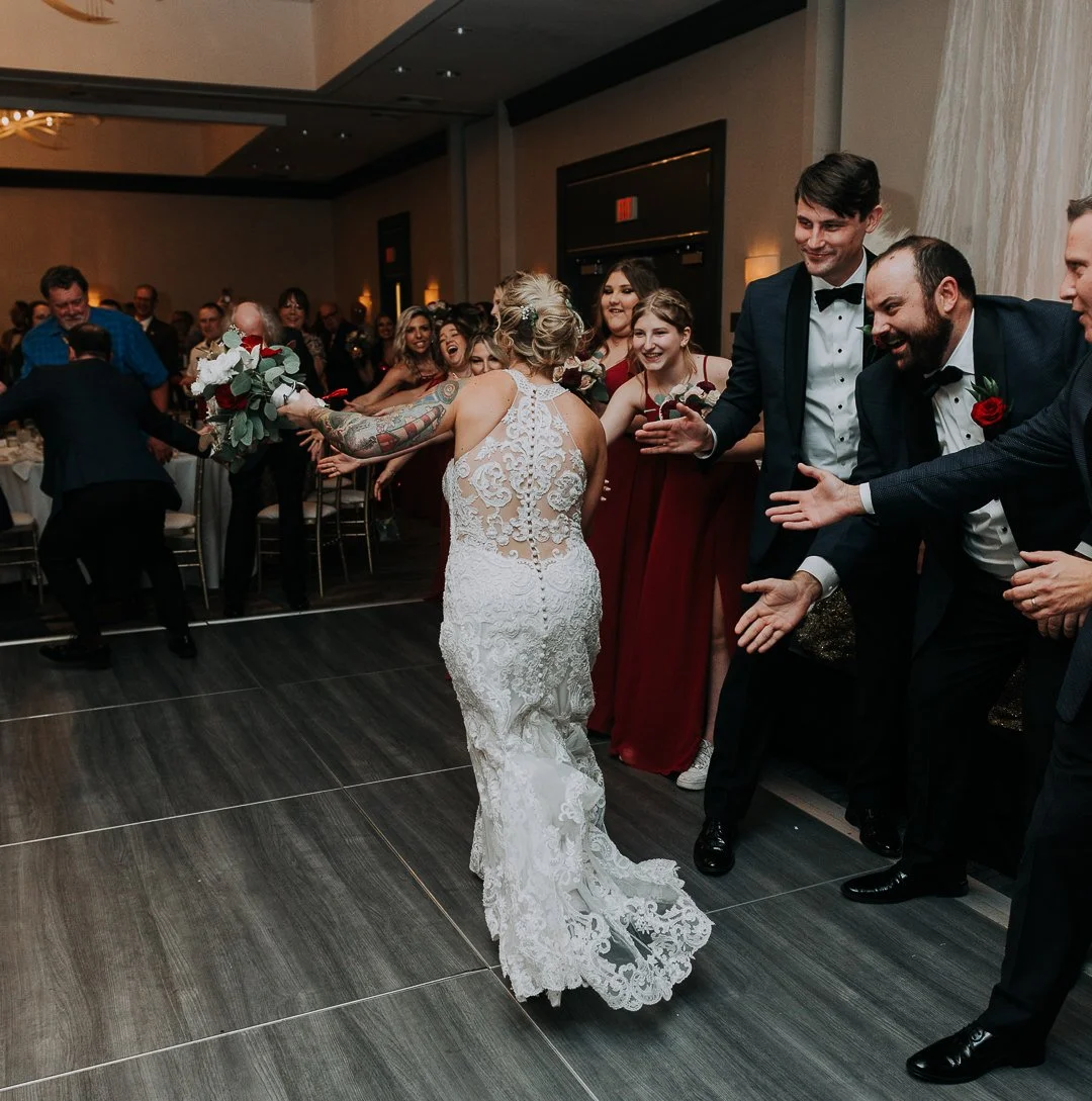 Bride in a lace wedding dress reaching out to guests at a wedding reception, with guests smiling and clapping.