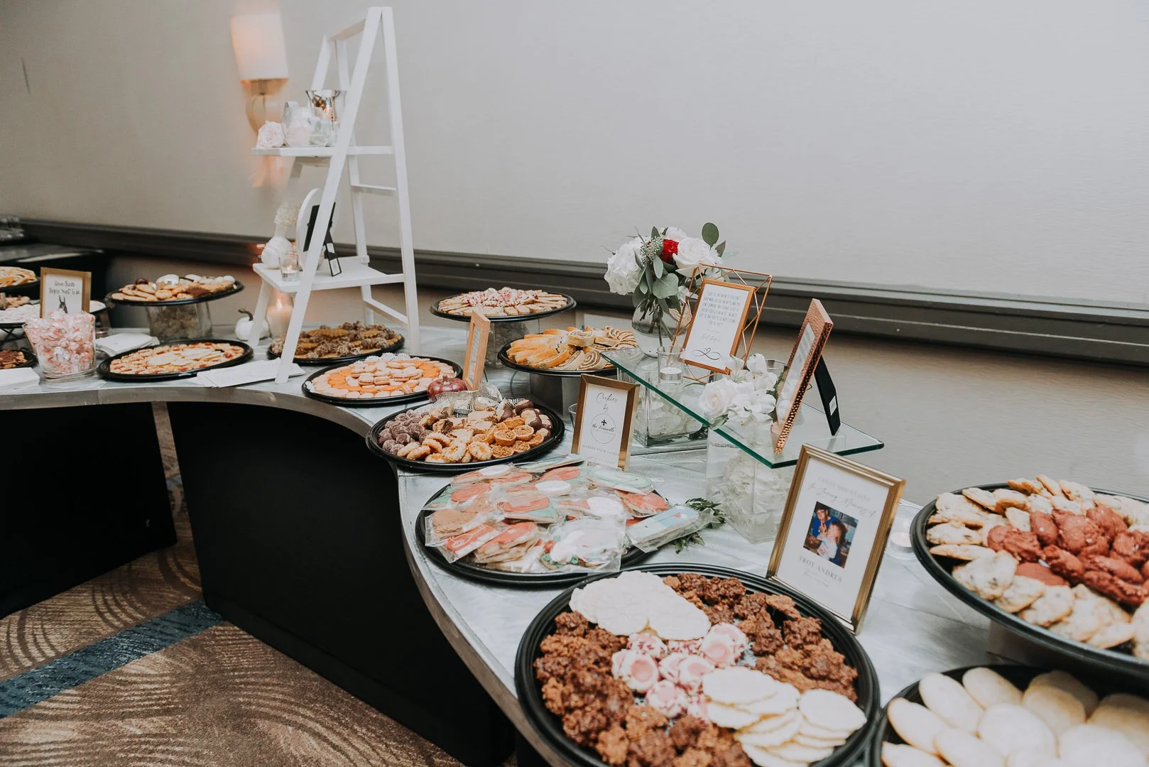 A dessert table featuring various cookies, pastries, and treats arranged on black trays. Decor includes framed photos, flowers, and small signs with descriptions.