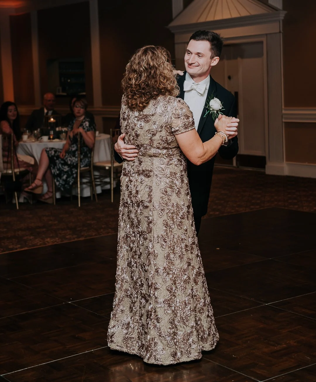 A young man in a tuxedo dances with an older woman in a long, patterned dress during a wedding reception, with guests seated at tables in the background.