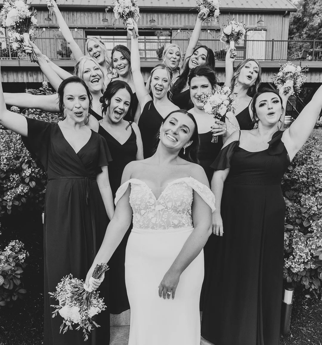 A bride and her bridesmaids celebrating outdoors, with the bride in a white dress holding a bouquet, and the bridesmaids in black dresses holding flowers, all smiling and cheering.