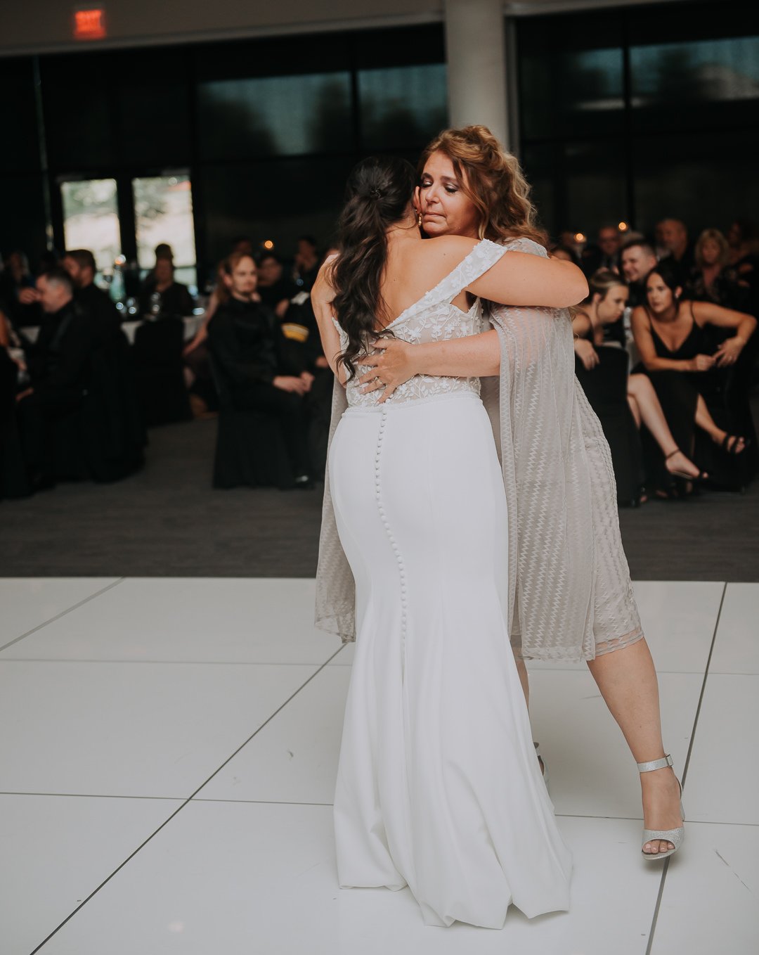 Two women hugging on a dance floor at a wedding reception, with guests seated at tables in the background.