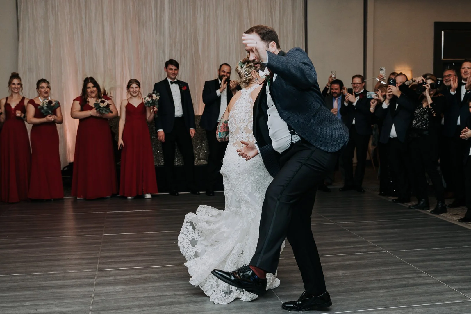 A groom in a dark suit and a bride in a white lace wedding dress dancing at their wedding reception, with guests standing in the background capturing the moment.