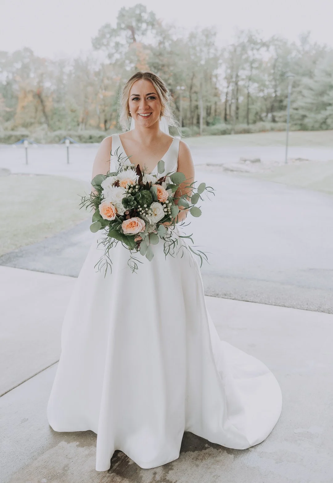 A smiling bride in a sleeveless white wedding gown holding a bouquet of roses, eucalyptus, and other flowers outdoors with trees in the background.