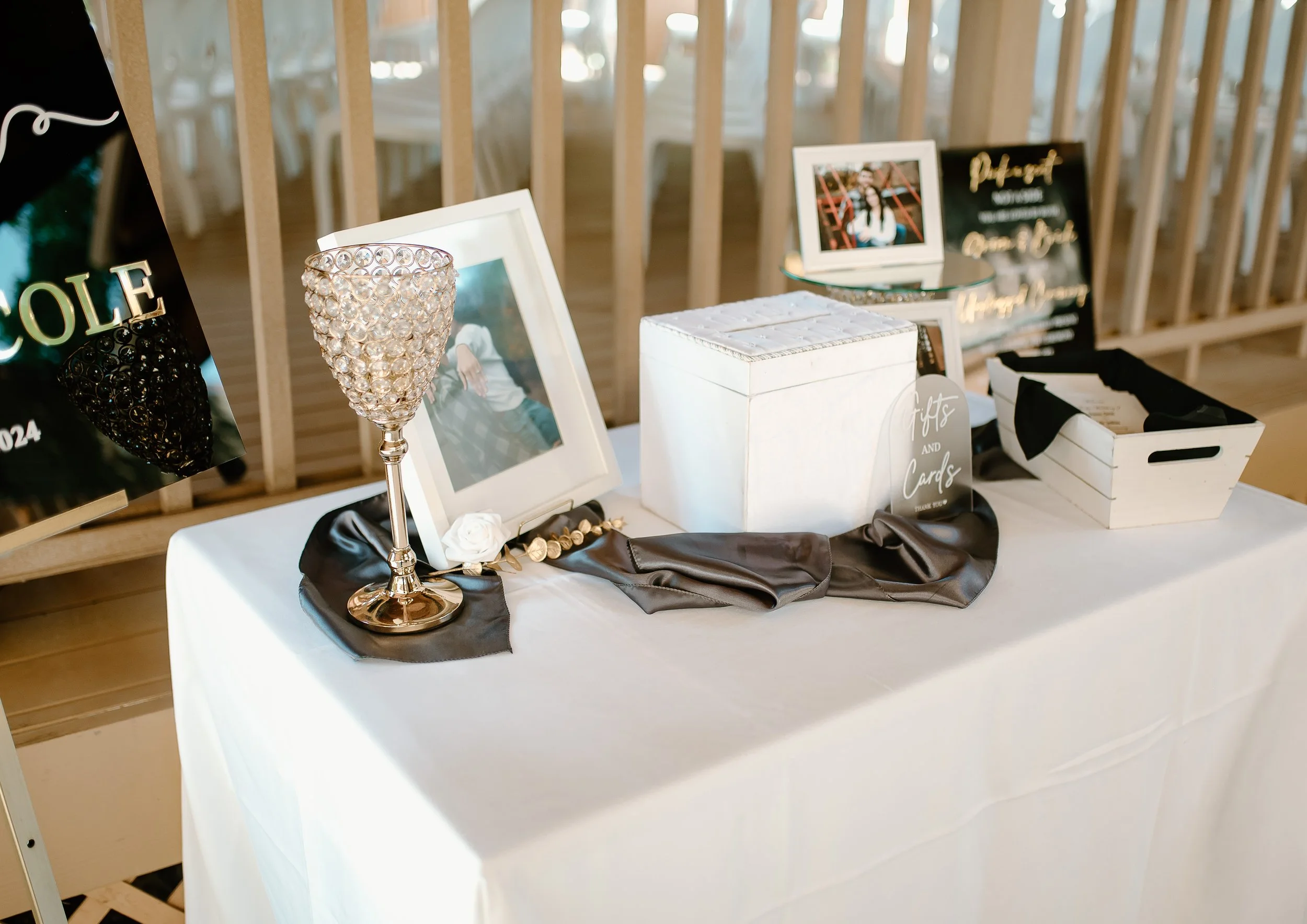 A wedding guestbook table with a white tablecloth, framed photo, decorative crystal goblet, white box, sign, and pictures, set against a wooden backdrop.
