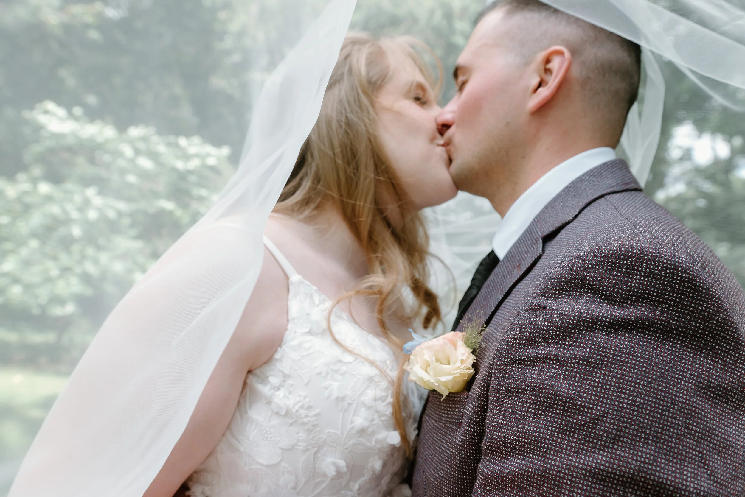 A bride and groom share a kiss under a sheer veil, outdoors with green foliage in the background.