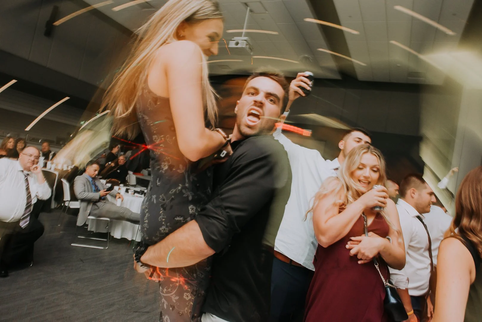 People dancing and celebrating at an indoor event, with some holding drinks and others using a phone, in a lively party atmosphere.