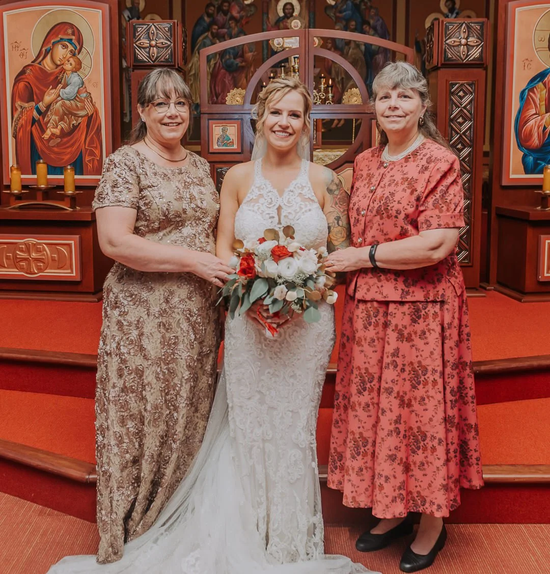A bride in a white lace wedding dress holding a bouquet stands between two women in a church. The woman on the left wears a gold patterned dress, and the woman on the right wears a red floral dress with black shoes. The background features religious 