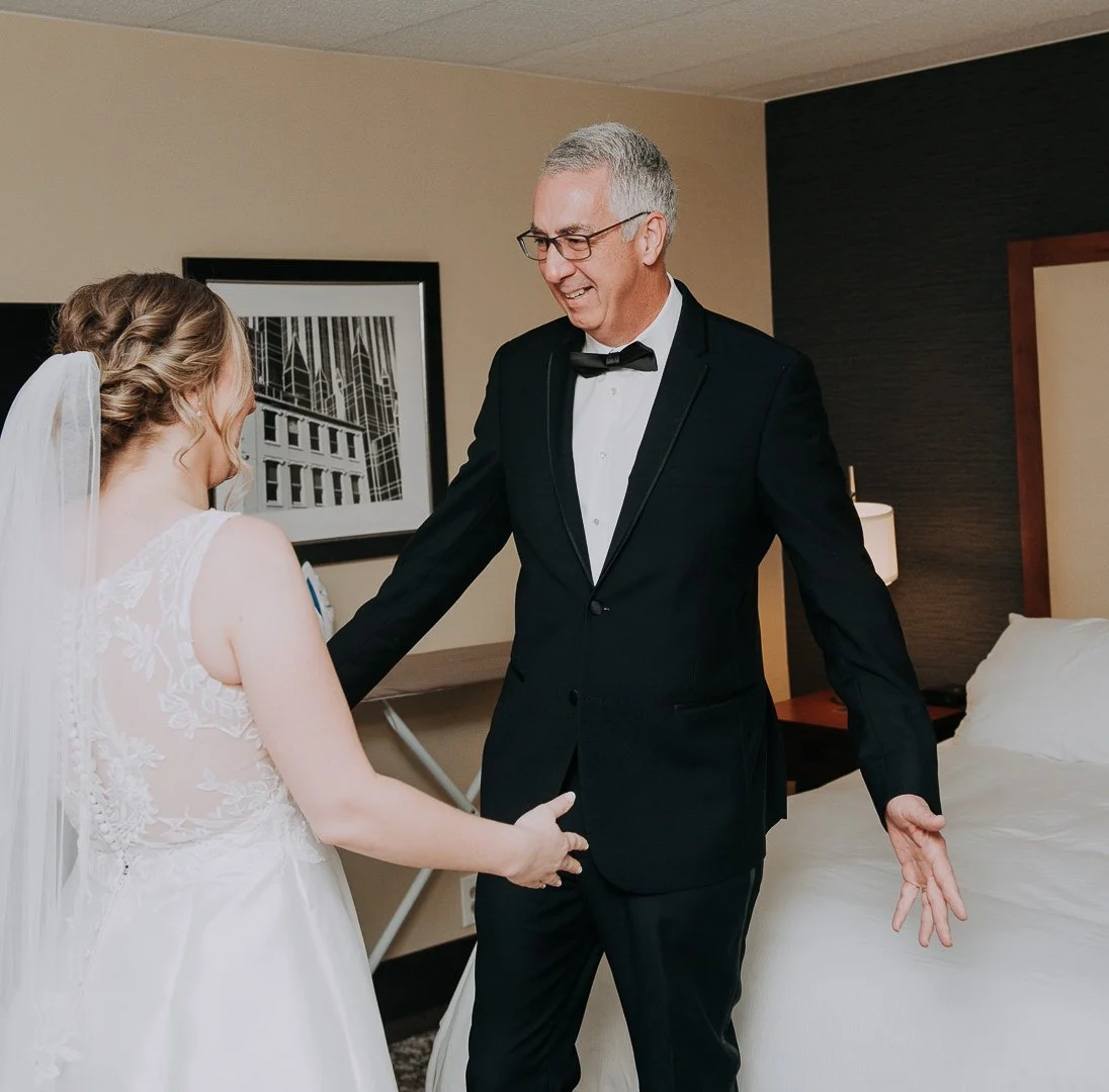 An older man in a tuxedo smiling and holding hands with a woman in a wedding dress inside a hotel room.