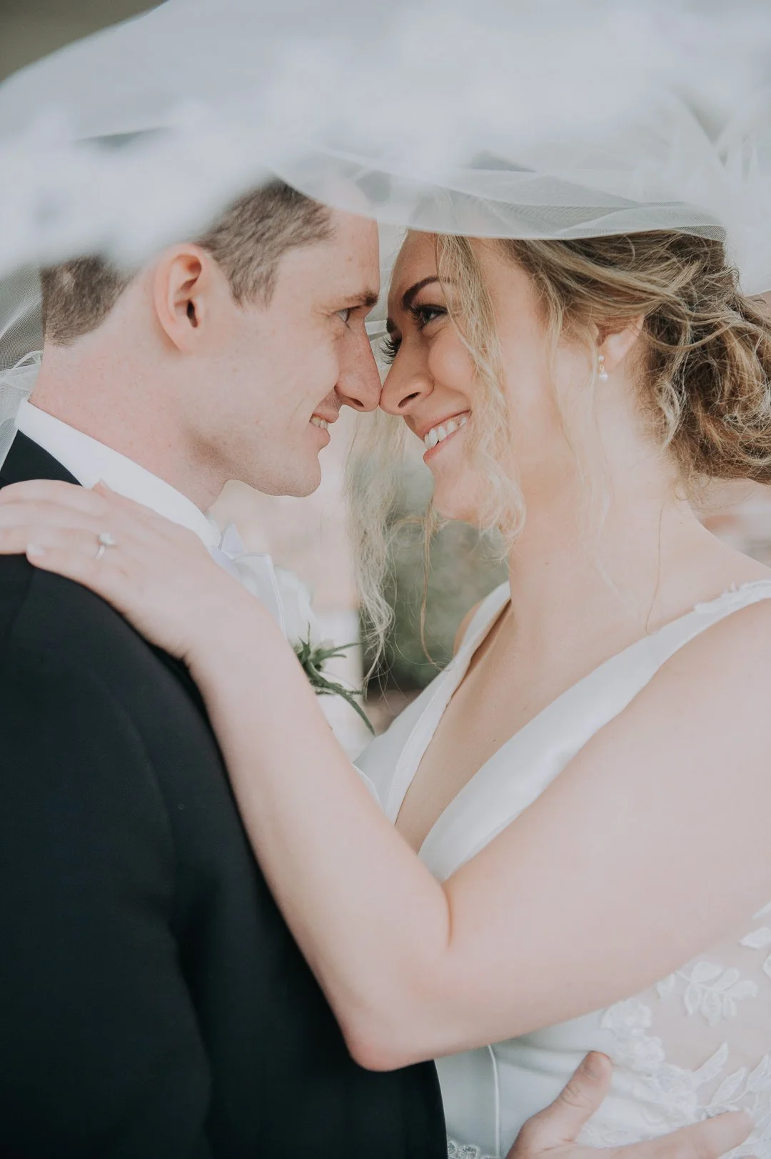 A bride and groom are close together, smiling, with their foreheads touching, under the bride's veil, during their wedding.