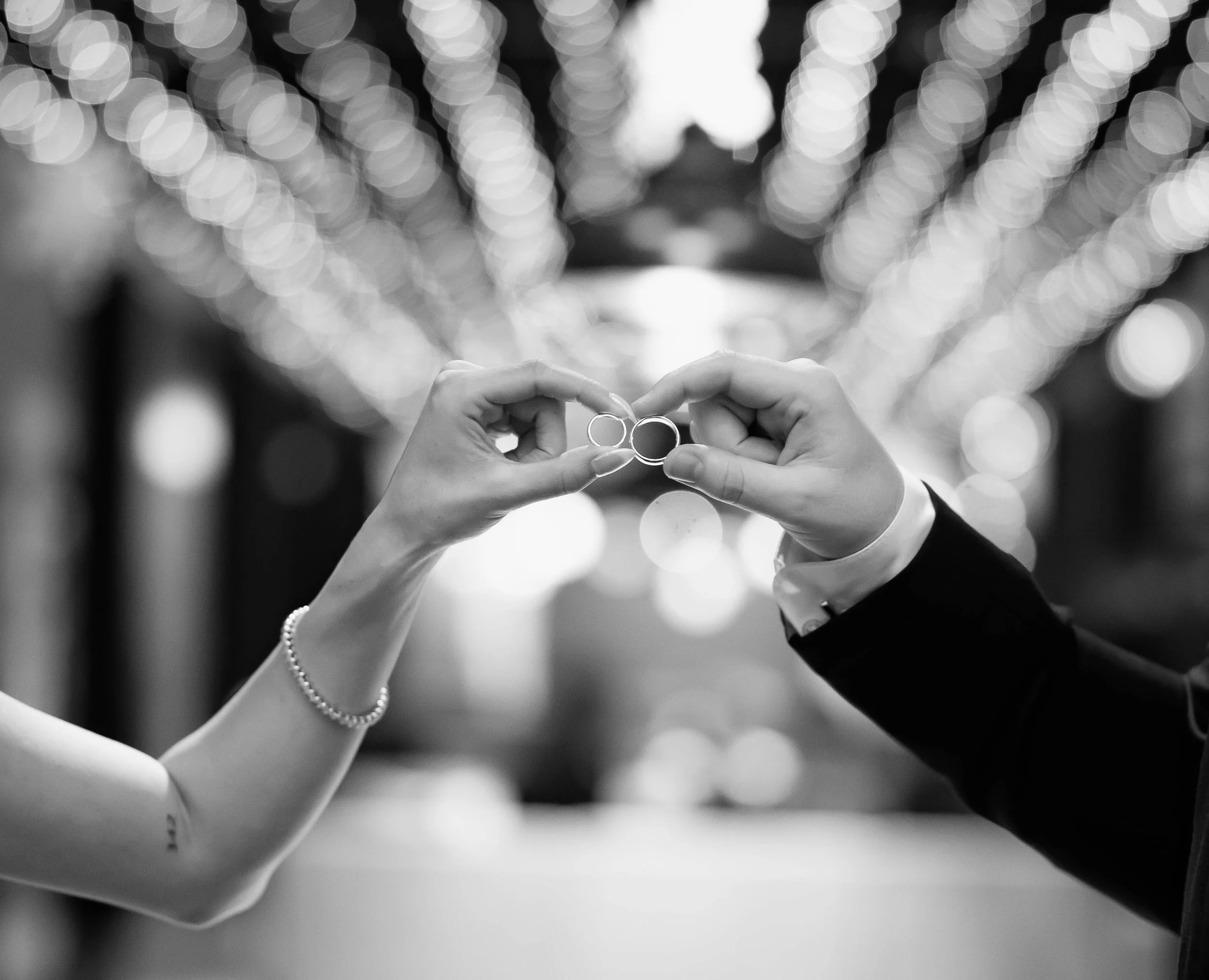 A black and white photo of a bride and groom holding wedding rings together, with the bride wearing a pearl bracelet and the groom in a suit, against a blurred background of lights.