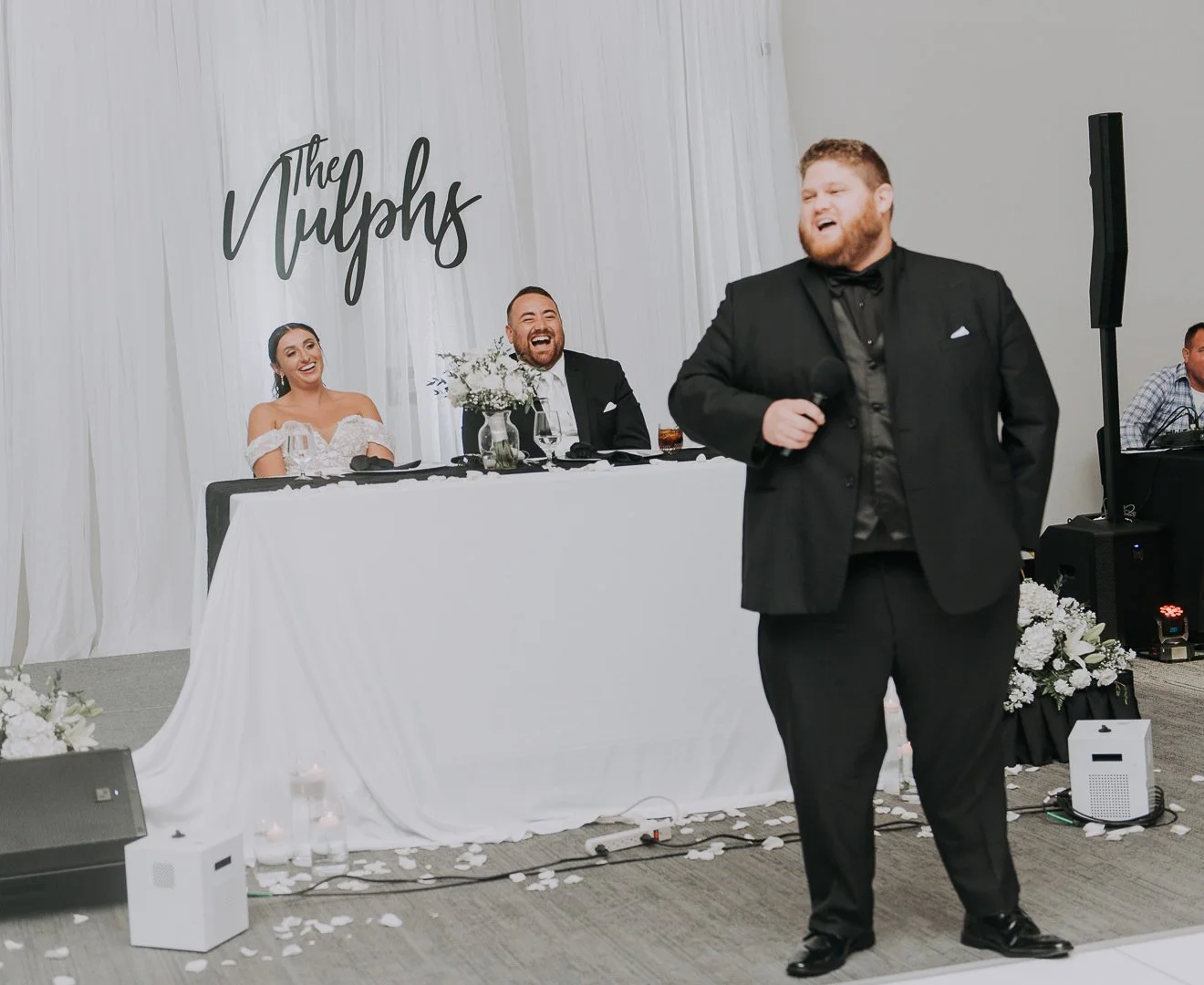 Man in a black tuxedo giving a speech at a wedding reception, with a bride and groom seated at a decorated head table behind him, and a backdrop with the text 'The Tulips'.