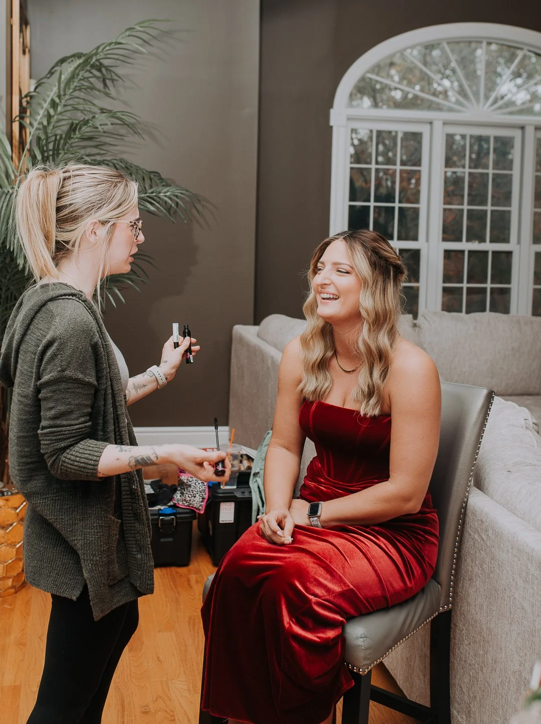 A woman in a red velvet dress sitting on a chair, laughing as a makeup artist applies makeup to her face in a home setting with a large window and houseplants in the background.