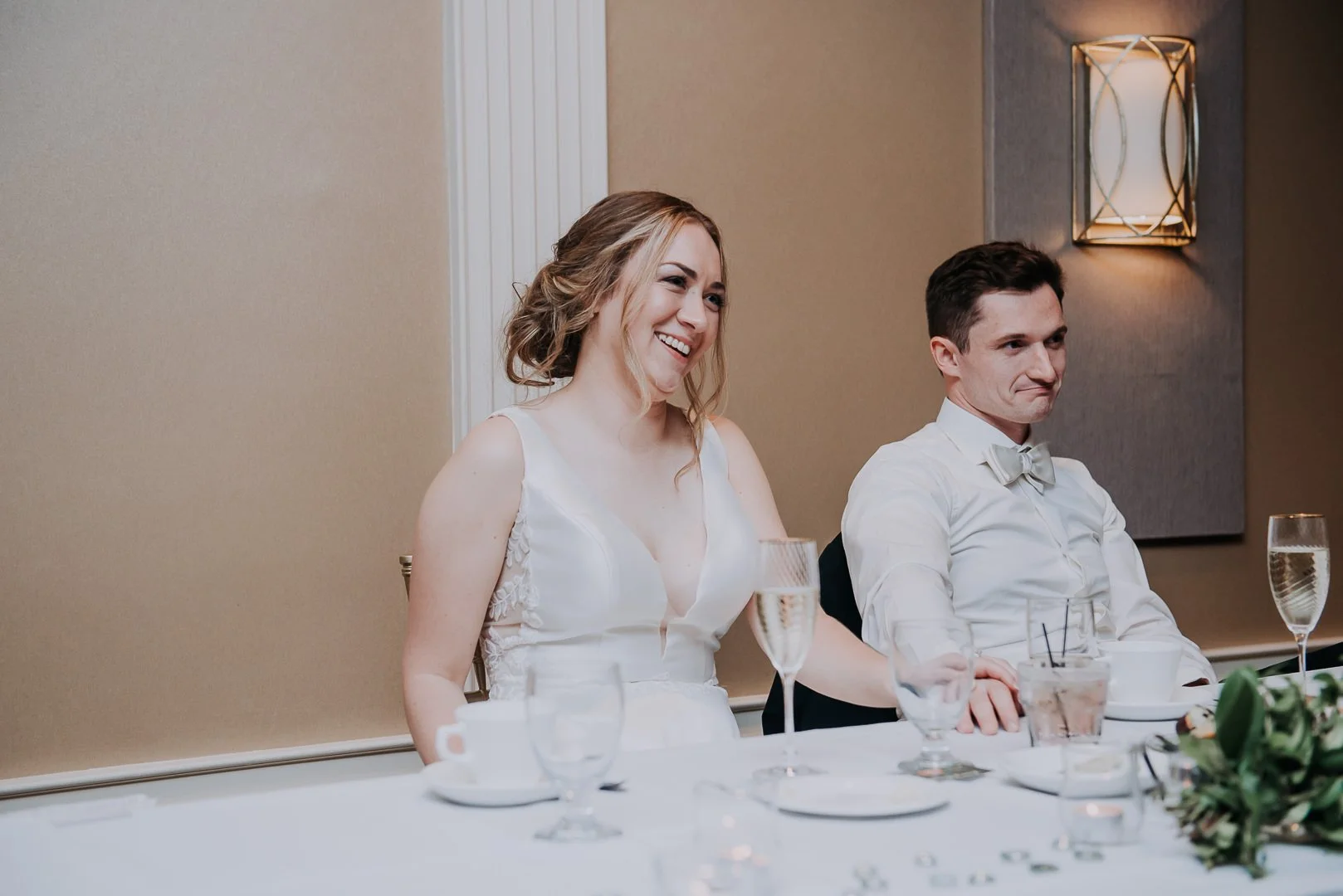 A woman in a white dress smiling and a man in a white shirt with a gray bow tie sitting at a table with glasses and flowers during a wedding reception.