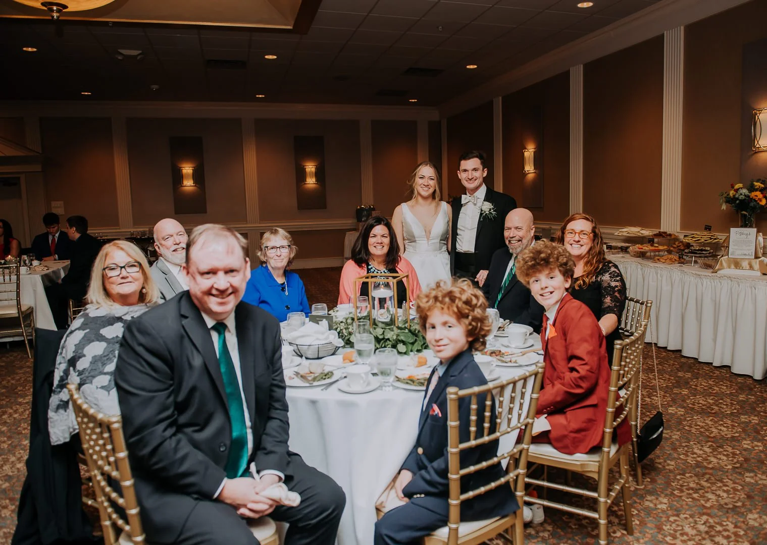 A group of people celebrating at a wedding reception, sitting around a round table with food and drinks, with a bride and groom standing behind them.