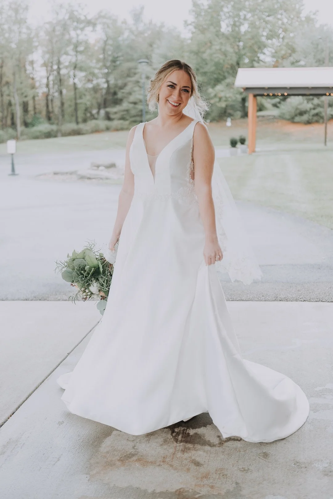 A bride in a white wedding dress holding a bouquet, smiling outdoors with trees and a pavilion in the background.