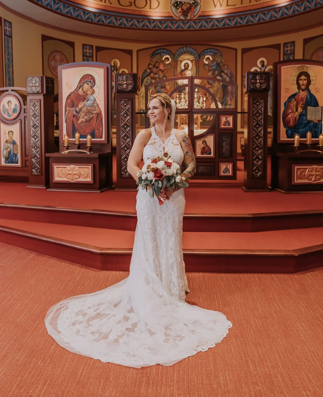 A bride standing inside a church in front of religious icons, holding a bouquet of white and red flowers, wearing a white lace wedding gown with a long train.