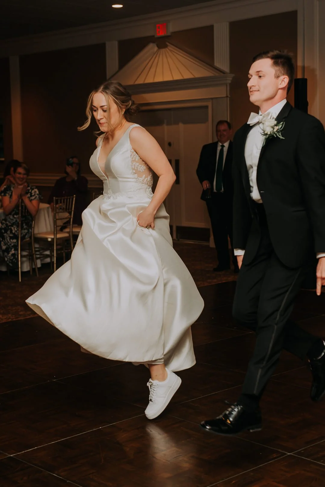 A bride in a white wedding dress and white sneakers dancing at her wedding reception, with a groom in a black tuxedo beside her and guests clapping in the background.