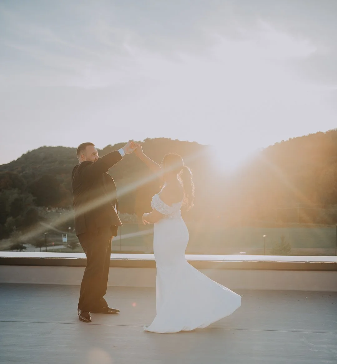 A newlywed couple dancing outdoors during sunset, with the groom in a suit and the bride in a white wedding dress, holding hands and spinning together.
