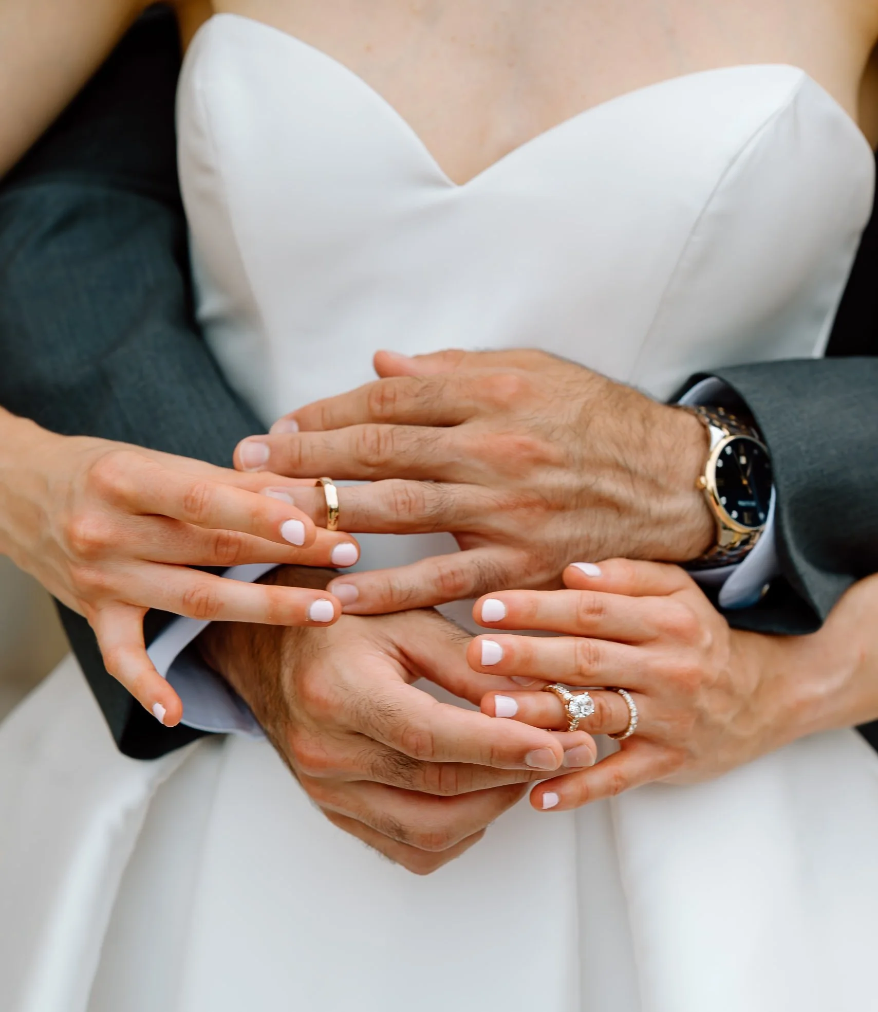 Close-up of a bride and groom holding hands, showing wedding rings, with the bride's white dress and the groom's suit.