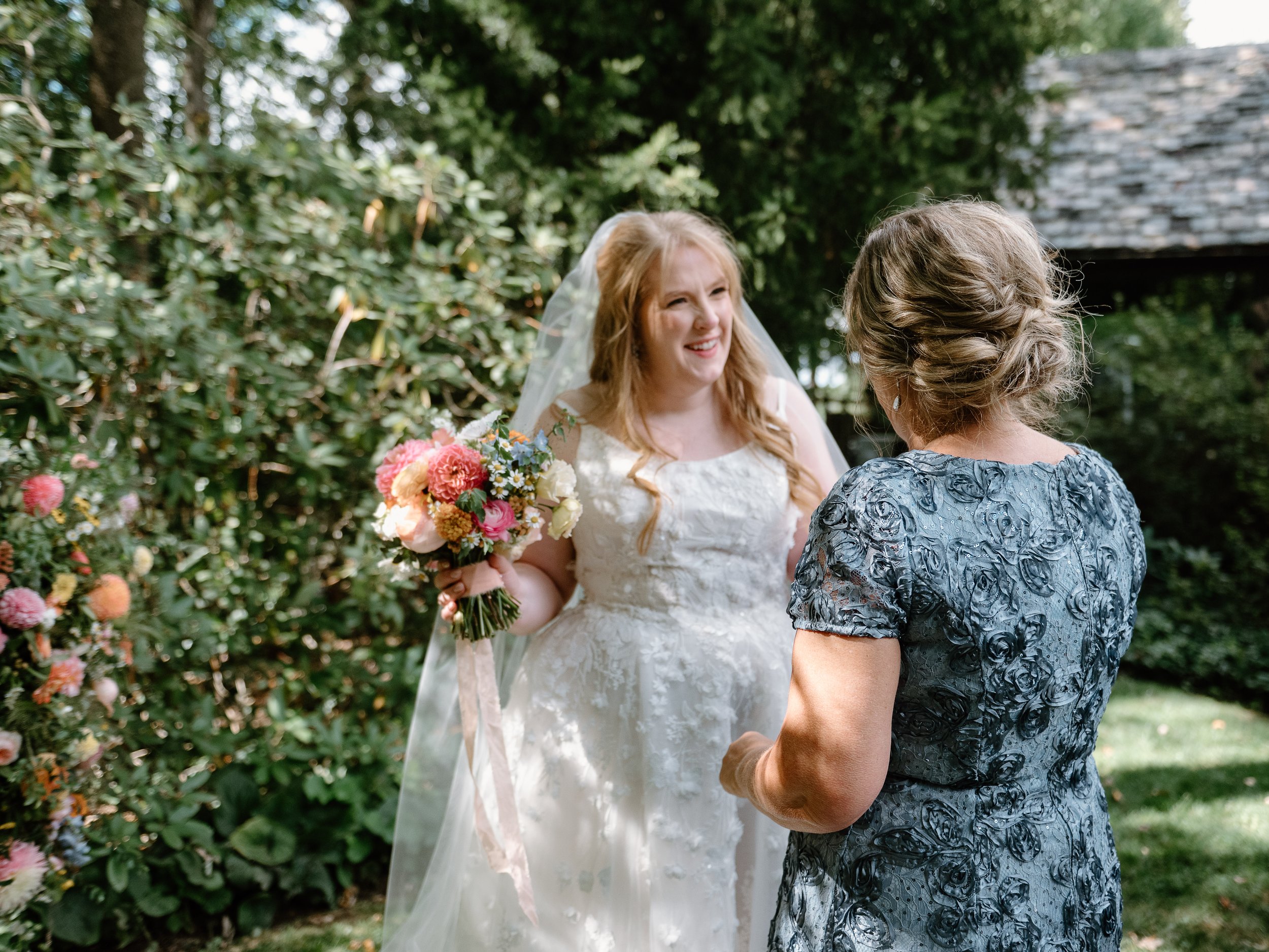 A bride with long red hair in a white wedding dress holding a bouquet, smiling at a woman in a blue patterned dress outdoors surrounded by greenery.